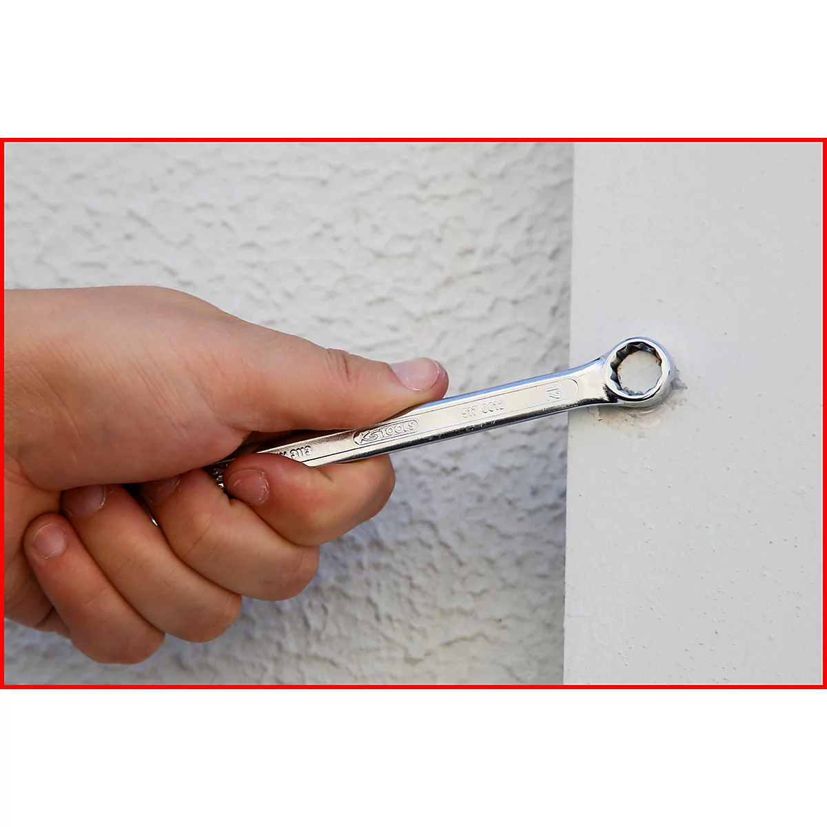 Eine Hand hält einen silbernen Ringschlüssel, der an eine weiße Wand gehalten wird. Der Hintergrund ist strukturiert.